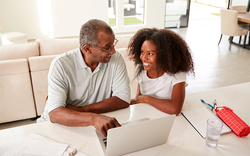 Smiling grandfather and granddaughter using a laptop at a table in a bright living space.
