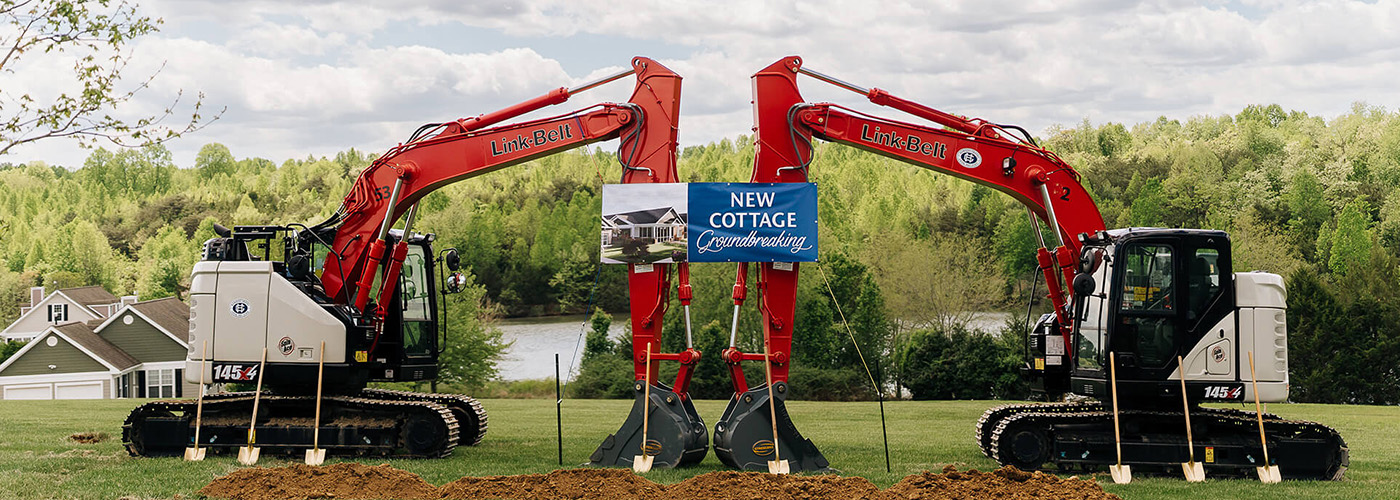 Two excavators at a groundbreaking ceremony for new living units near a wooded area.