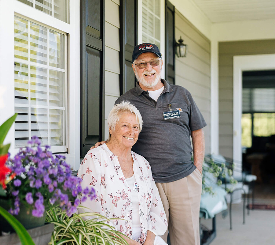 Smiling elderly couple sitting on a porch with flowers at their senior living community.