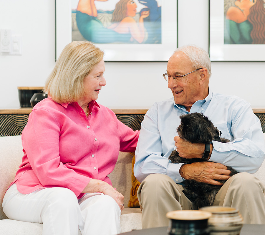 Older couple smiling, seated on a sofa, with a small black dog.