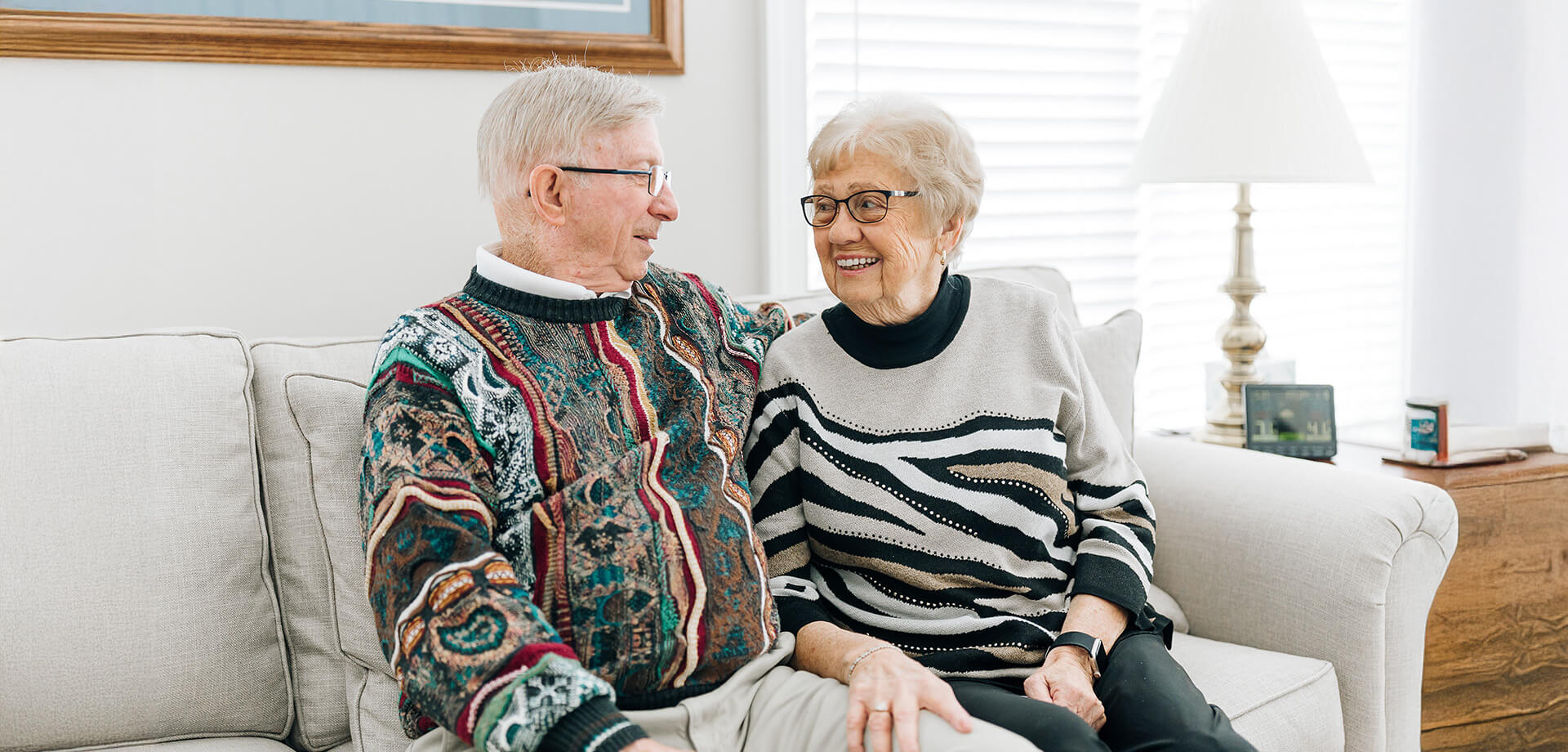 Elderly couple smiling and sitting together on a beige couch in a well-lit living room.