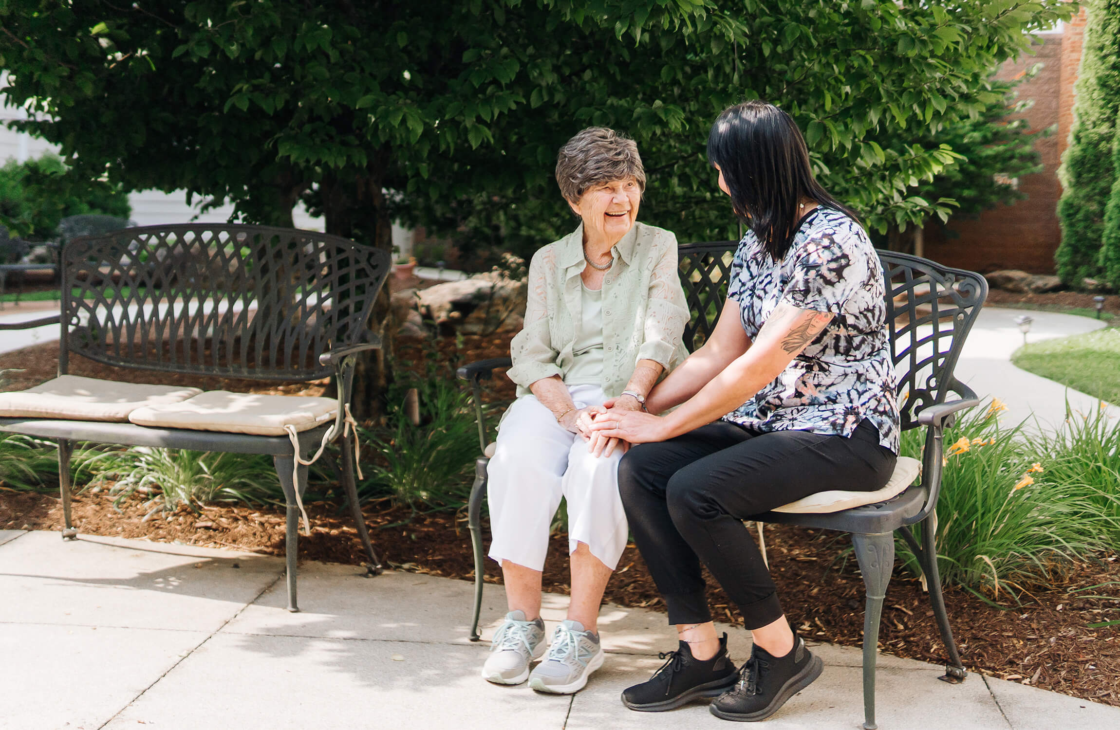 Older woman and caregiver sitting on a bench in a garden, sharing a moment of laughter.