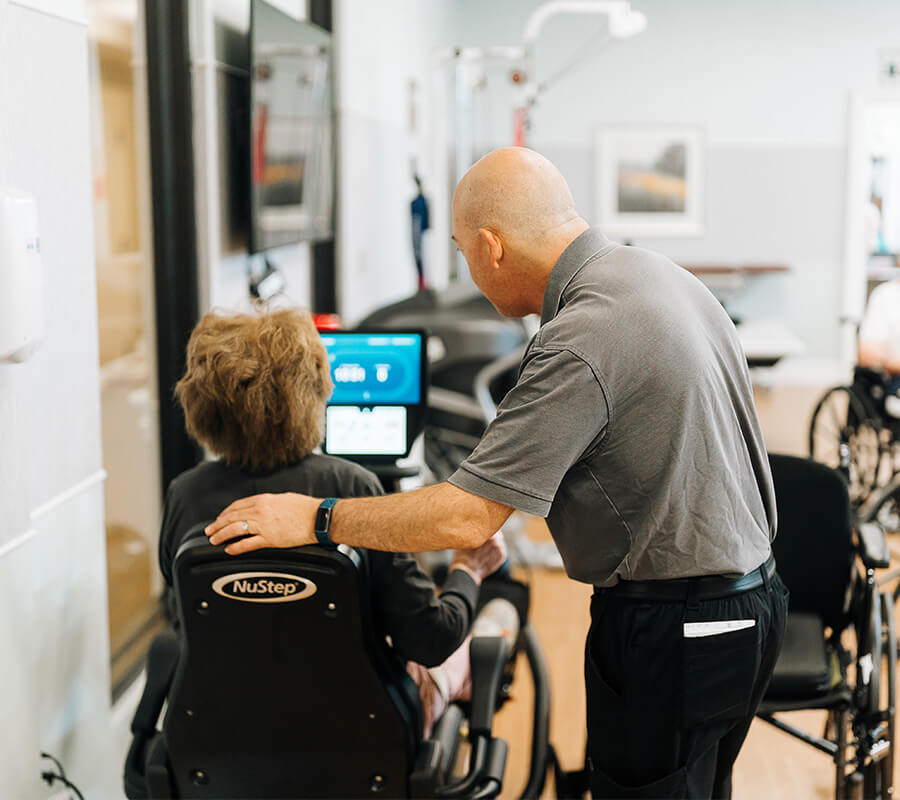 Instructor assisting senior on exercise machine in community room.