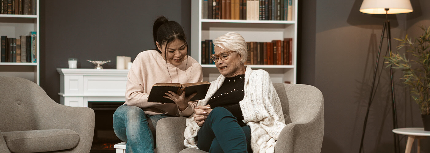 A young woman reads to an elderly woman in a cozy living room setting.