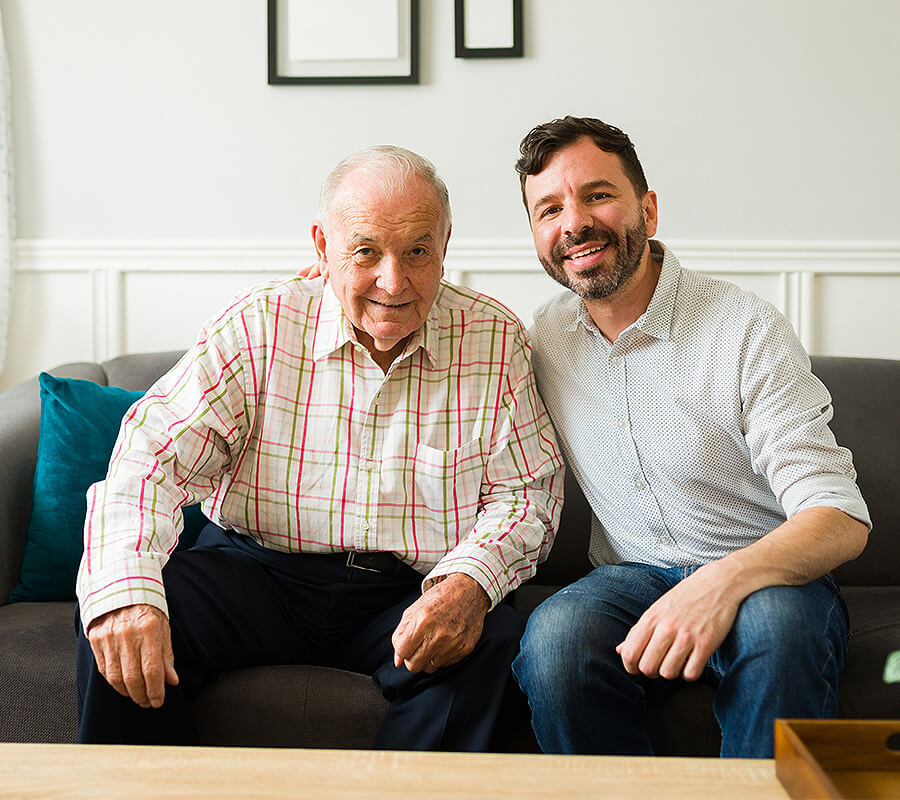 Older adult and younger man sitting on a couch in a brightly lit living space.