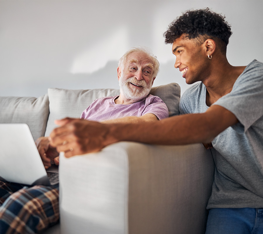 Elderly man and young man smiling and using a laptop together on a couch.