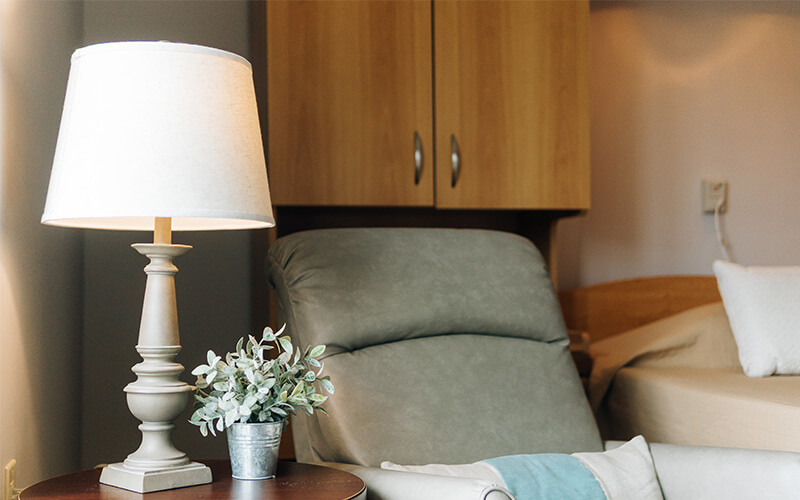Cozy unit interior with a lamp, armchair, and bed in a senior living community.