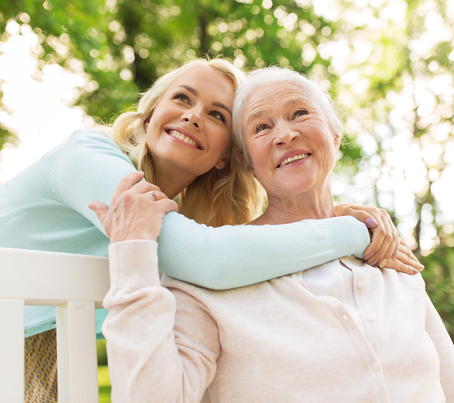 Two women smiling happily on a park bench surrounded by lush green trees.