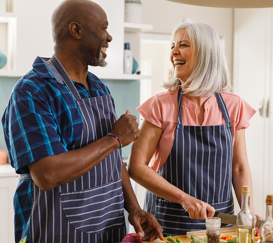 Two seniors cooking together wearing aprons and smiling in a kitchen.