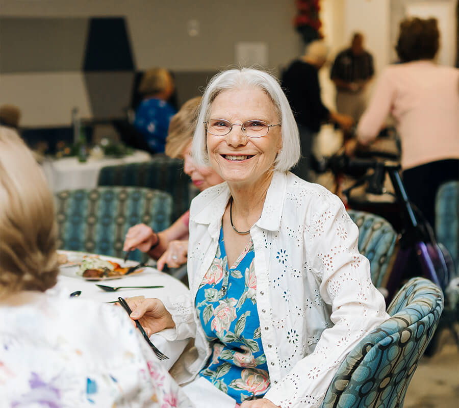 Elderly woman smiling in dining area, surrounded by people in a senior living unit.