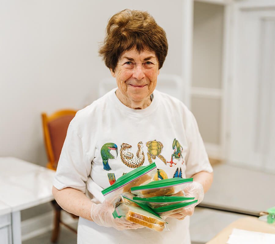 Elderly woman in white shirt holding bagged sandwiches in a bright room.
