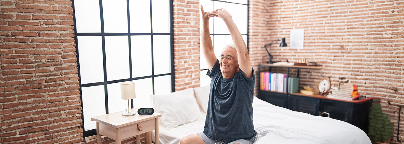 Senior man stretches on bed in sunlit brick-walled bedroom with a peaceful smile.