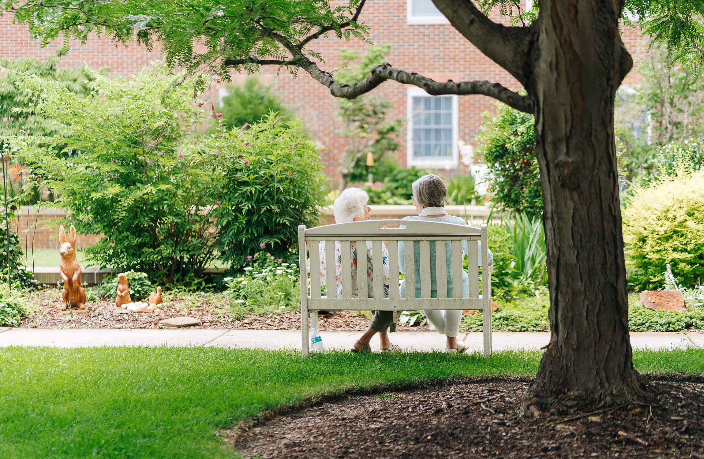 Two elderly women sitting on a bench in a lush garden, with a brick building in the background.