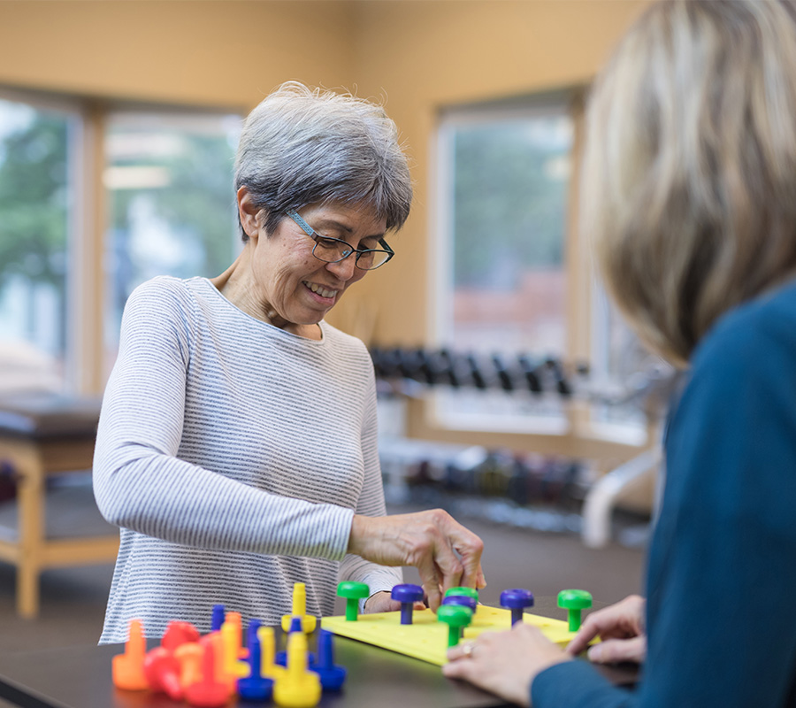 Smiling elderly woman engaged in colorful peg board activity with a therapist.