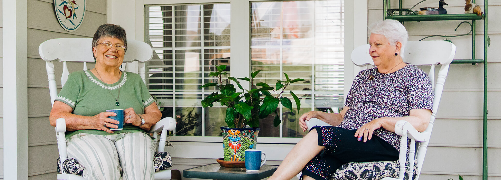 Two women sitting on a porch with rocking chairs and plants.