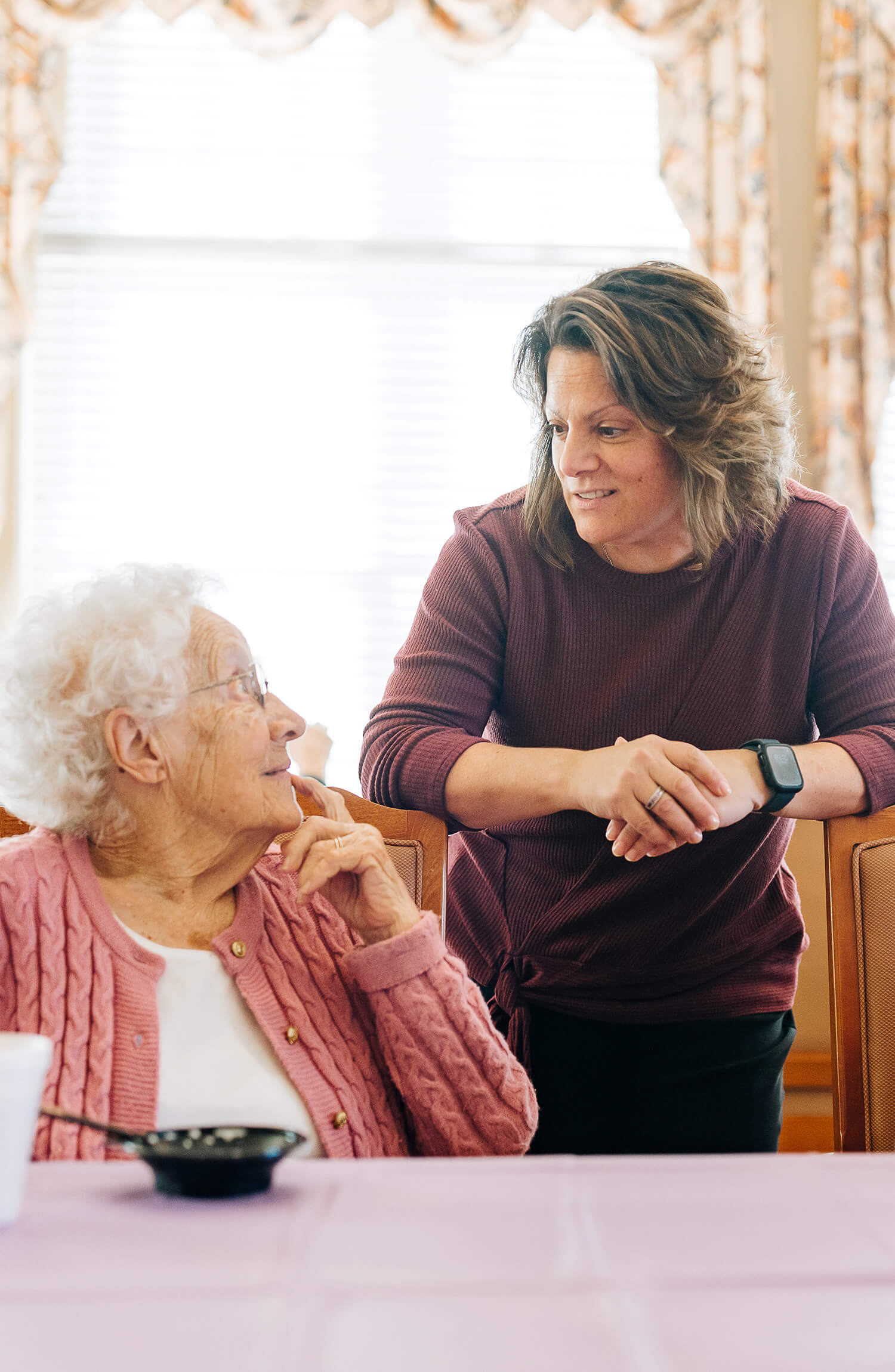 Elderly woman in pink sweater talks with staff member in a cozy living unit.