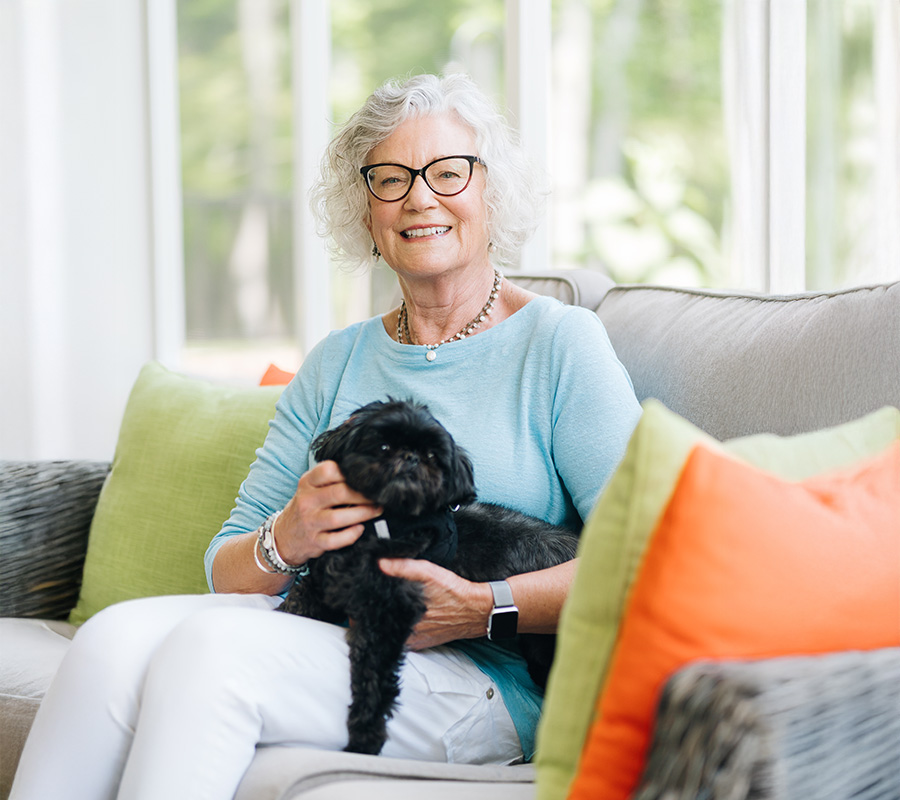 Elderly woman with glasses smiling and holding a small dog in a brightly lit room.