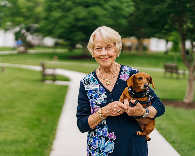 Elderly woman holding a dachshund on a scenic path in a senior community.