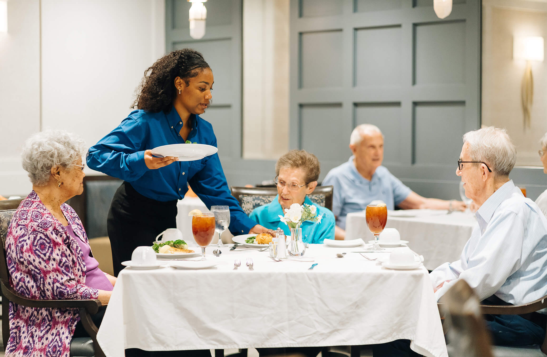 Residents in a dining area being served by a staff member in a senior living facility.