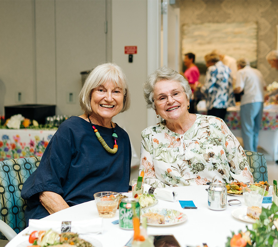 Two smiling women seated at a table in a senior community dining area.