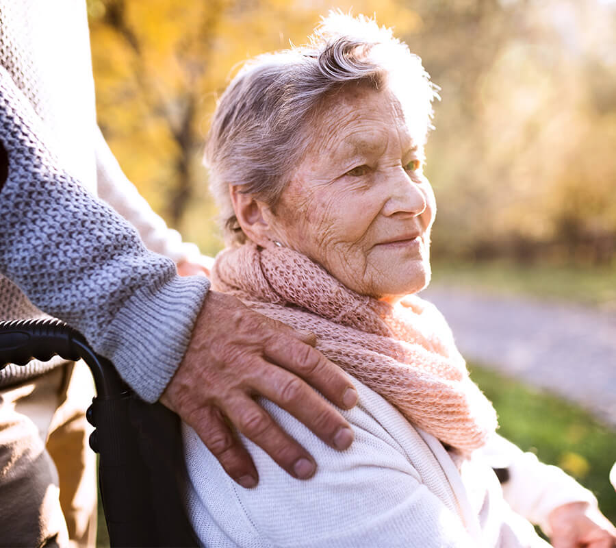Elderly woman in wheelchair outdoors, assisted by caregiver, in sunny autumn setting.