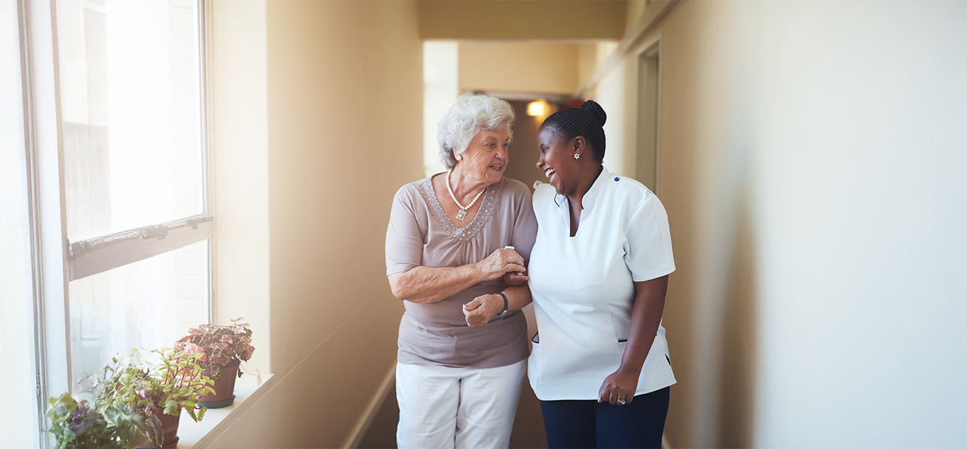 Elderly resident walking with caregiver in brightly lit hallway.