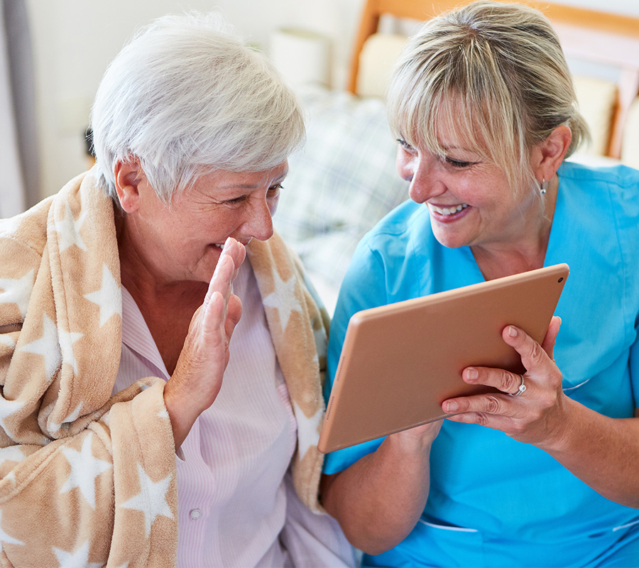 Senior woman in a robe waves at a tablet held by a caregiver in blue scrubs.