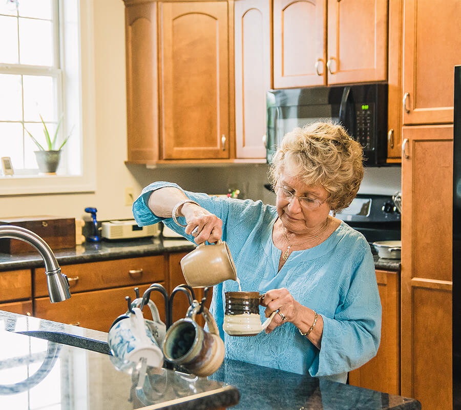Senior woman in blue blouse pouring coffee in kitchen at a living community unit.