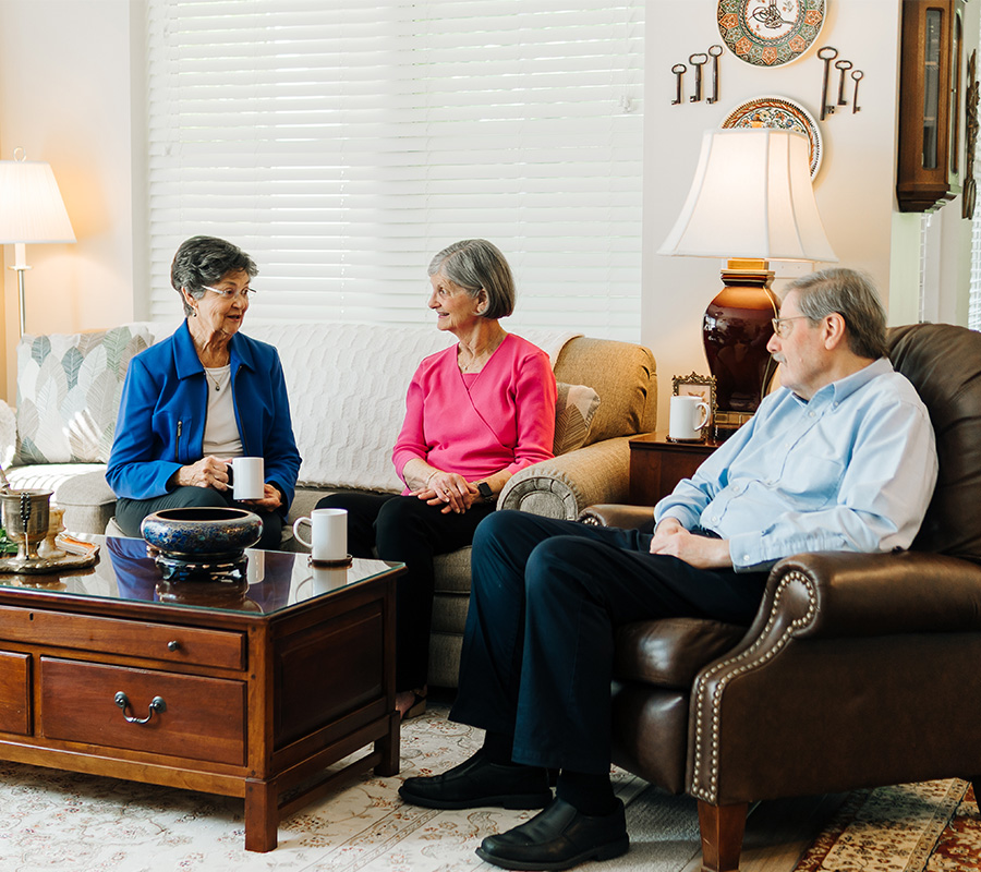 Three seniors enjoy a conversation in a cozy, well-lit living room.