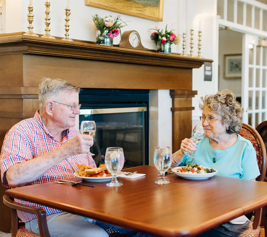Senior couple dining at a table in a cozy room with a fireplace.
