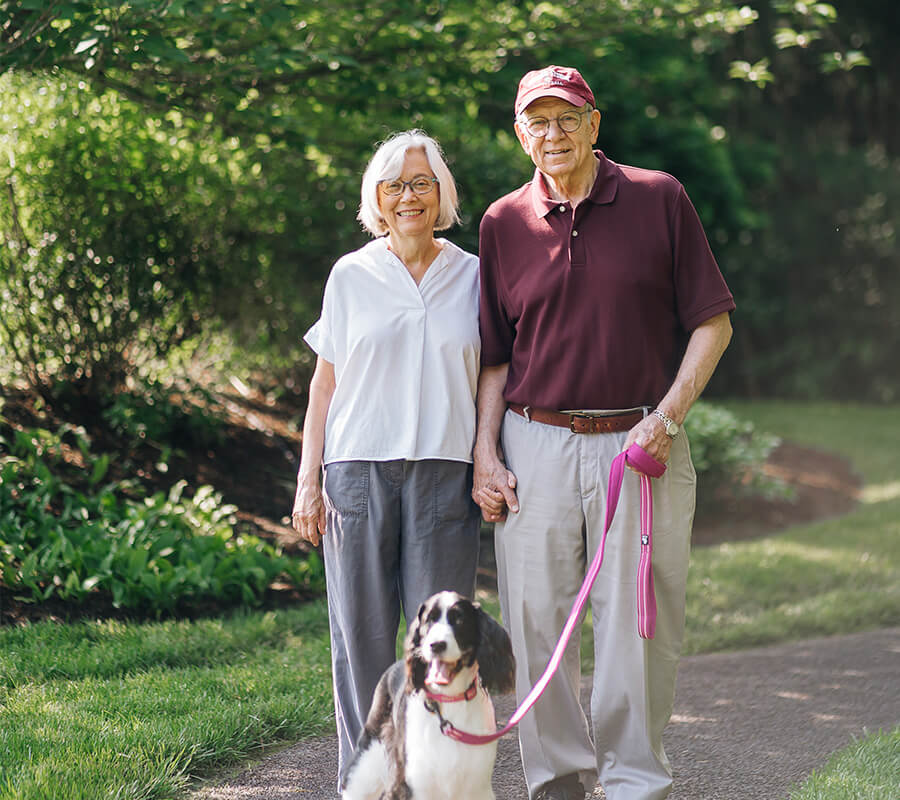 Senior couple walking their dog on a garden path, holding hands and smiling.