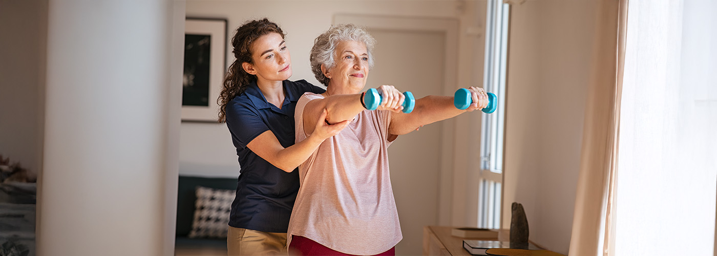 Elderly woman exercising with dumbbells guided by a caregiver in a bright unit.