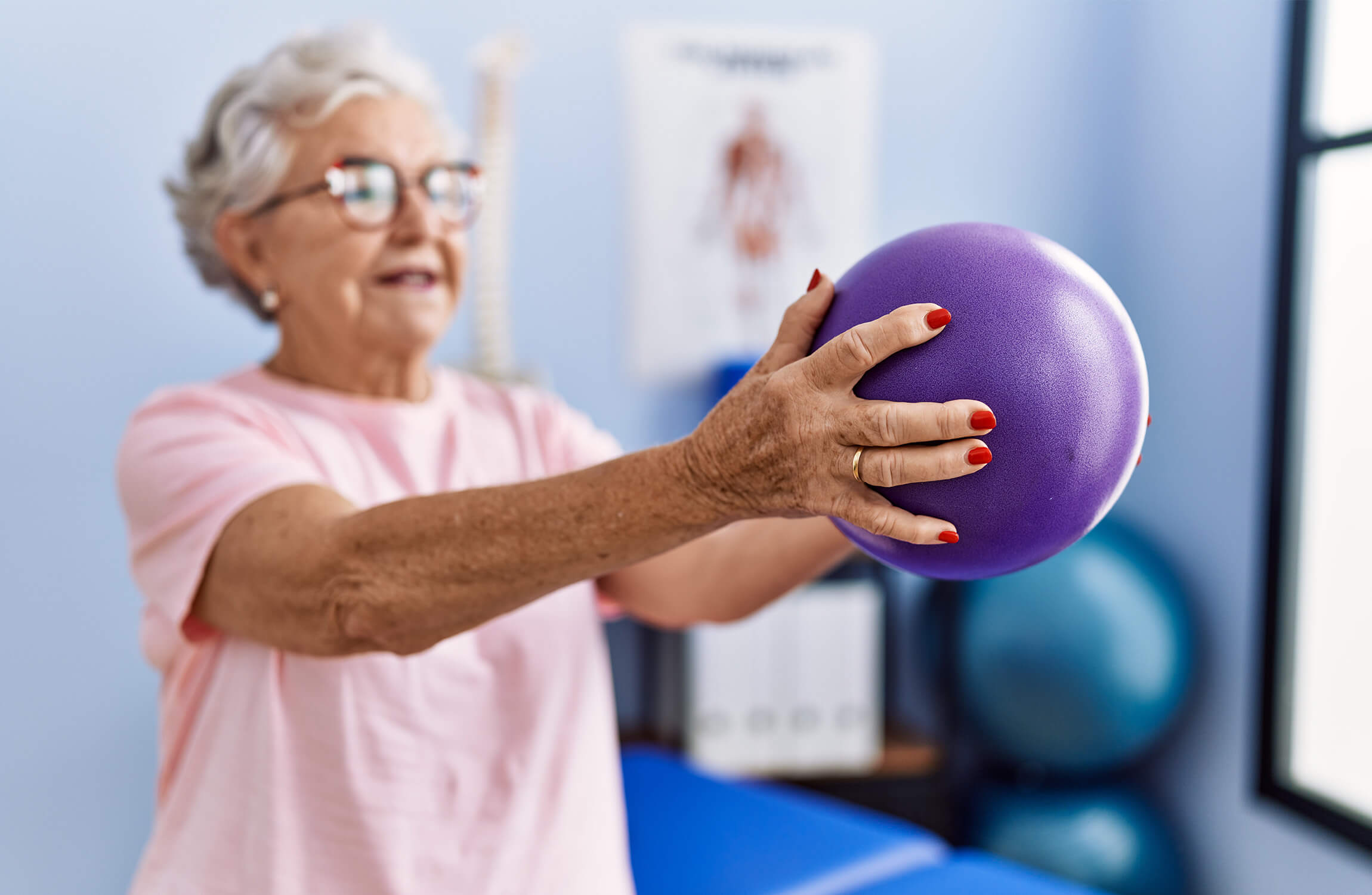 Senior woman in pink shirt exercising with a purple ball indoors.