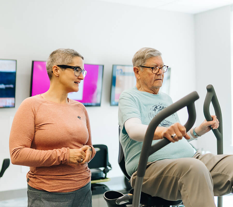 An elderly man exercises on a bike with a trainer in a fitness room.