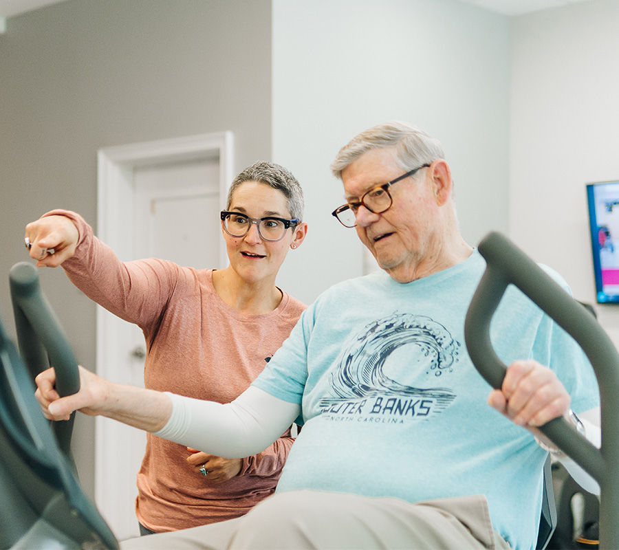 Senior man on exercise bike with trainer assisting in a fitness room.