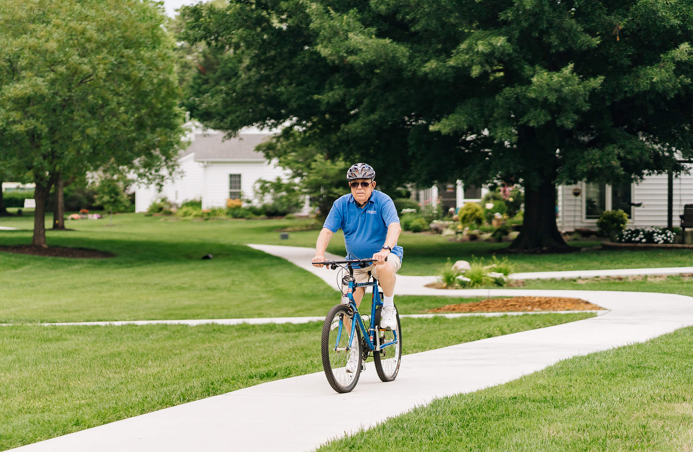 Senior man biking on a path through a green landscape with trees and units in the background.