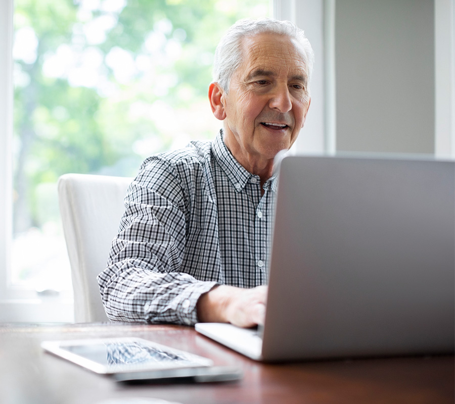 Senior man using a laptop at a table with a tablet nearby.