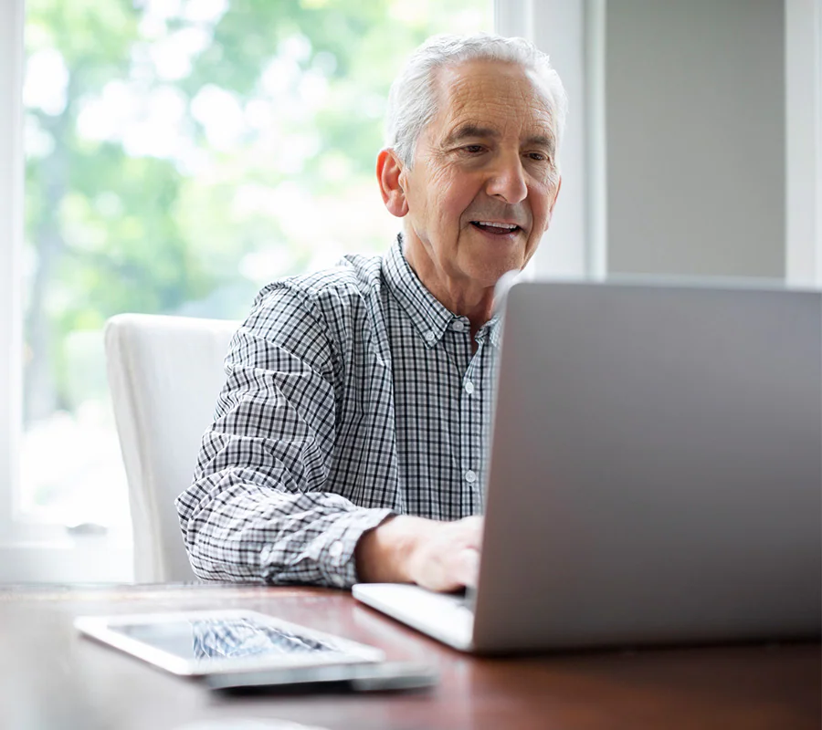 Senior man using a laptop at a table with a tablet nearby.