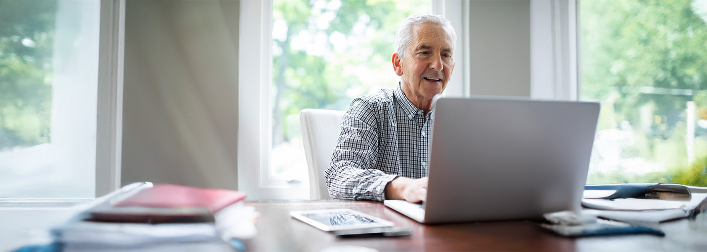 Senior man smiling while using a laptop at a desk with large windows.