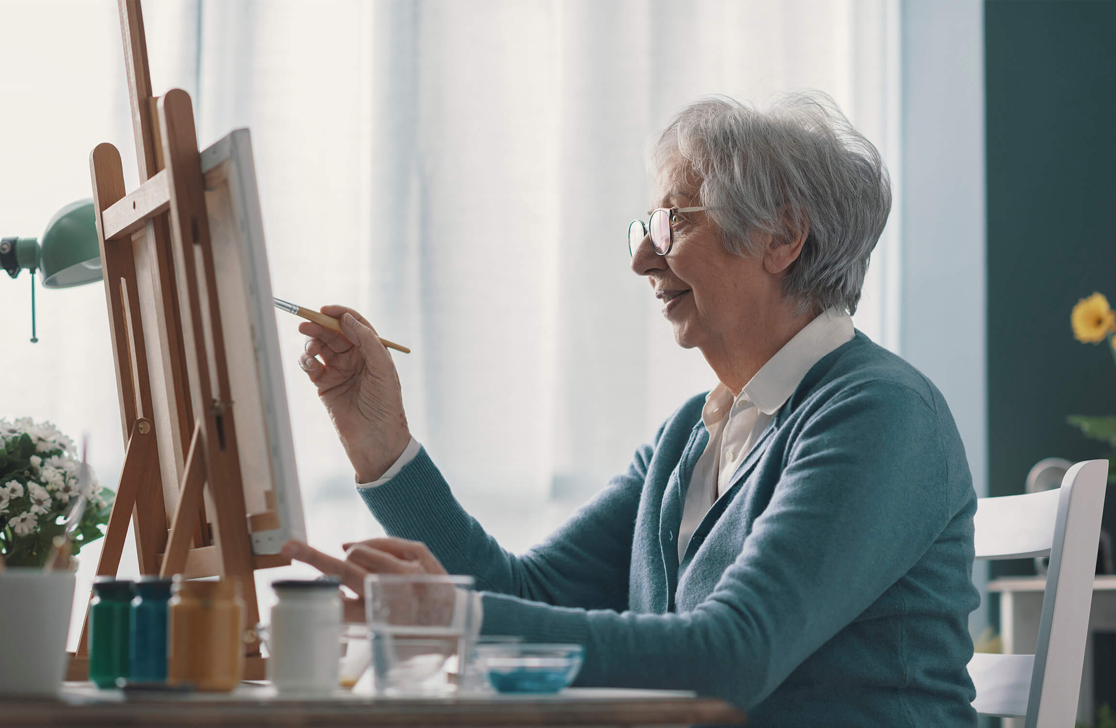 Elderly woman painting at an easel in a well-lit room.