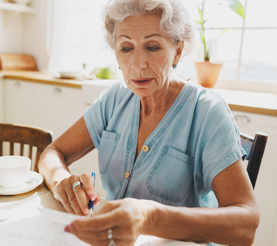 Senior woman in blue shirt reviewing documents at a kitchen table.
