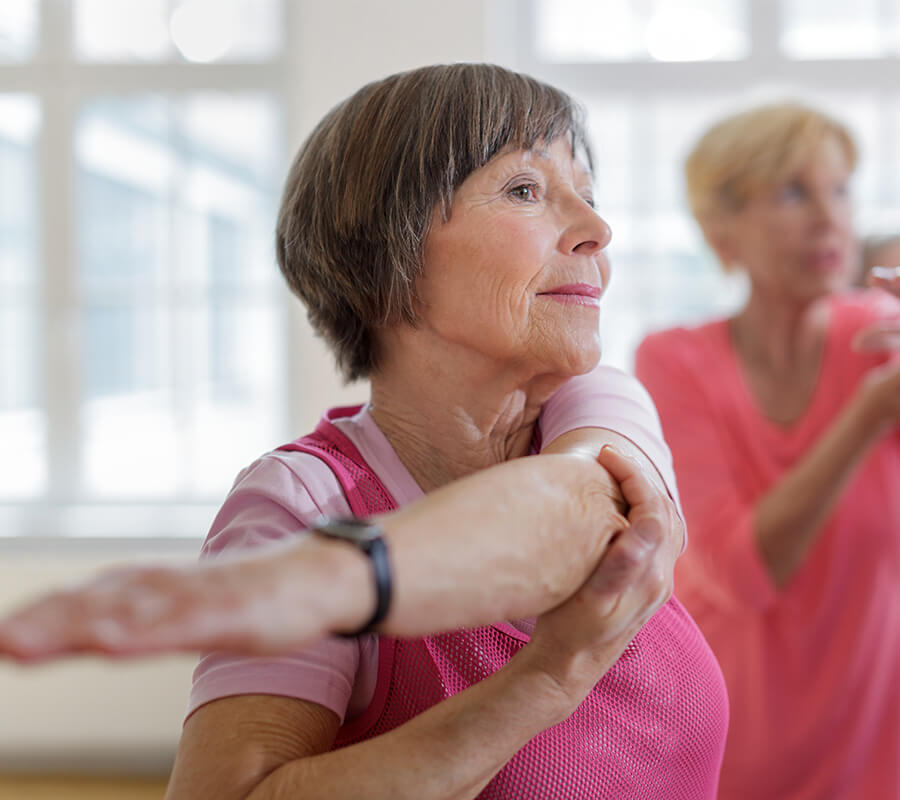 Older adult woman in a pink shirt stretching in a bright room with others.