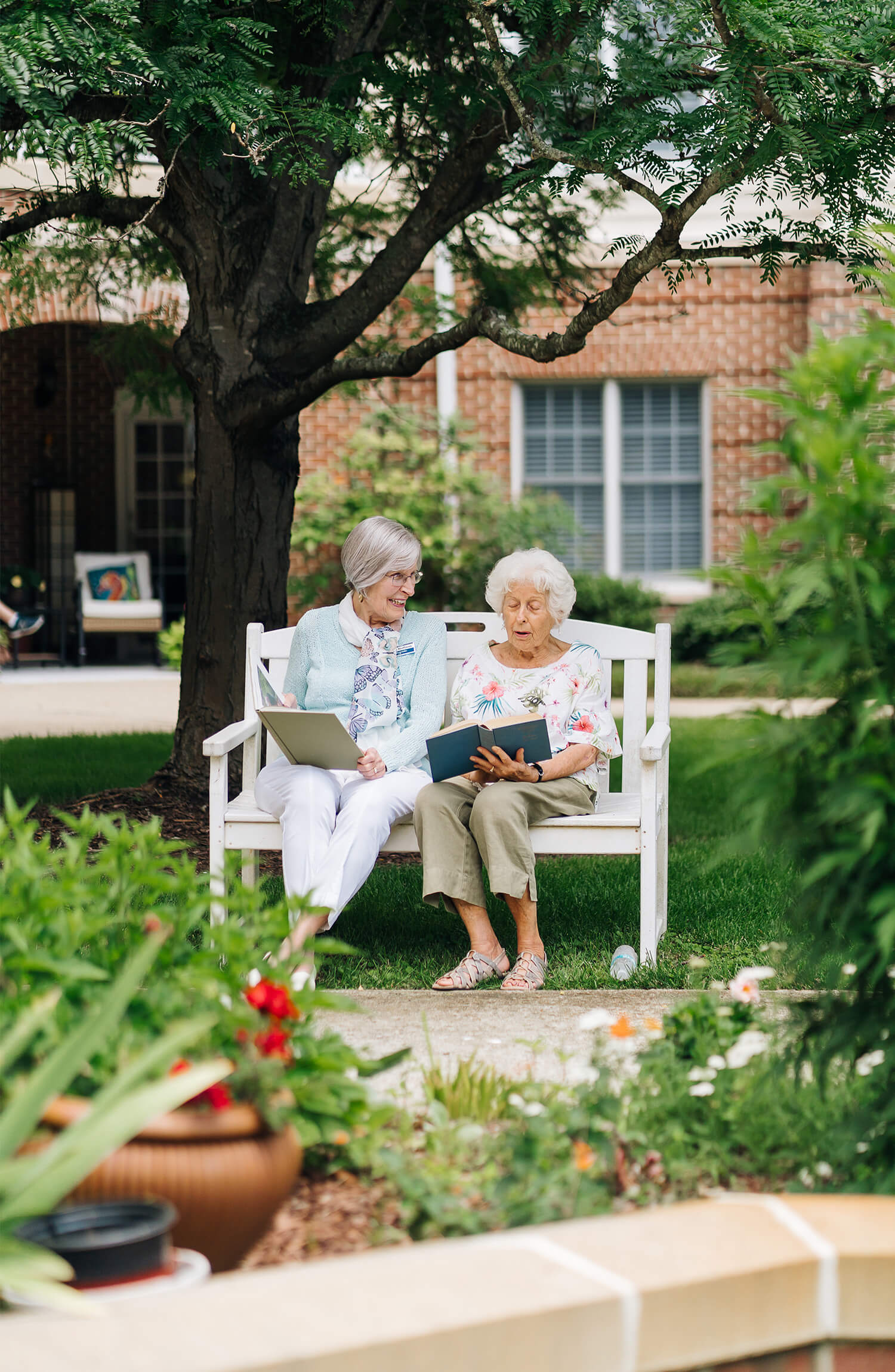 Two senior women read books on a bench in a garden at a senior living community.