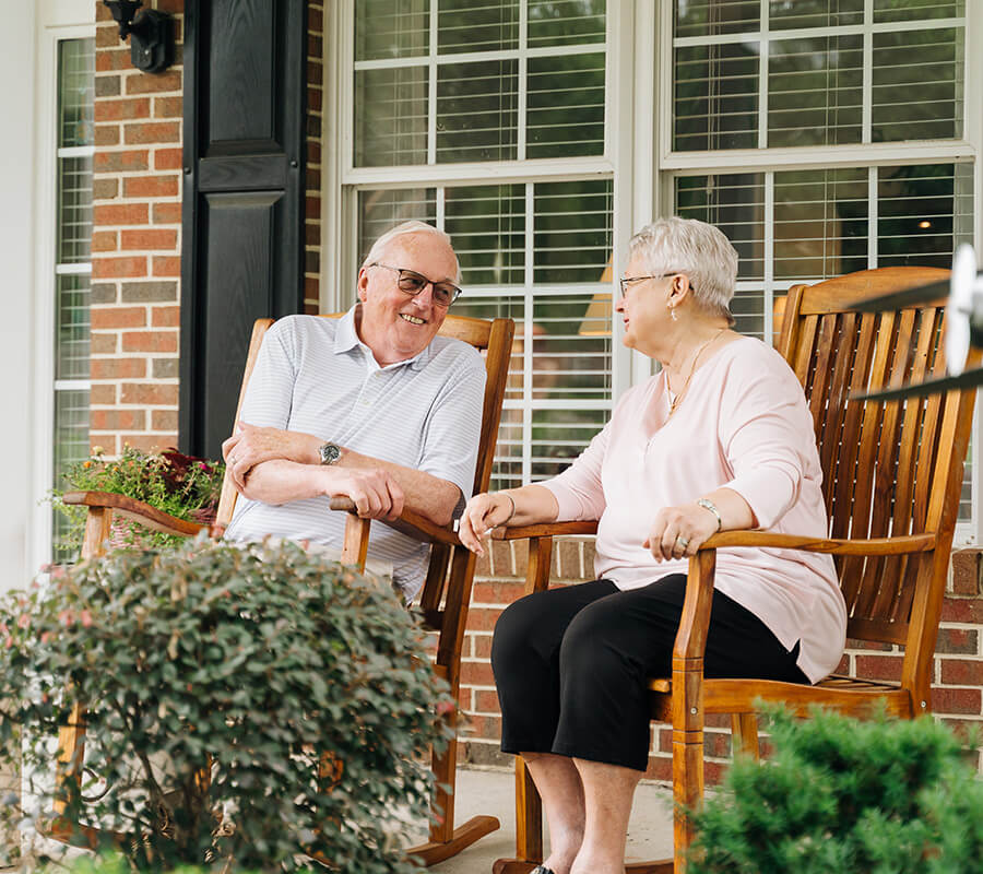 Two seniors happily chatting on a porch in wooden rocking chairs.