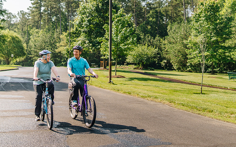 Seniors Enjoying a Bike Ride Two seniors cycling on a paved trail amidst greenery in a residential community.