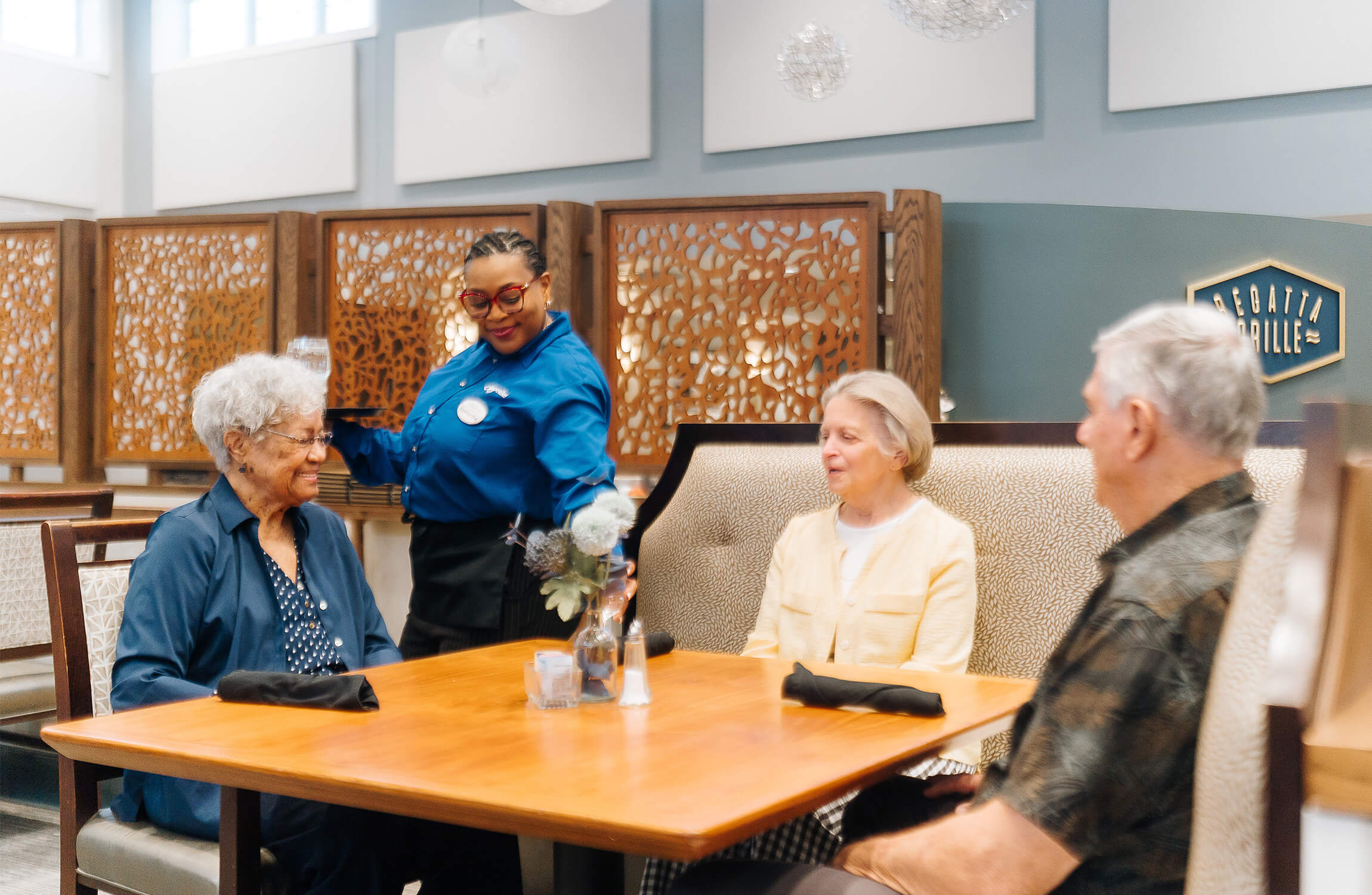 Seniors dining with a staff member serving water in a community dining area.