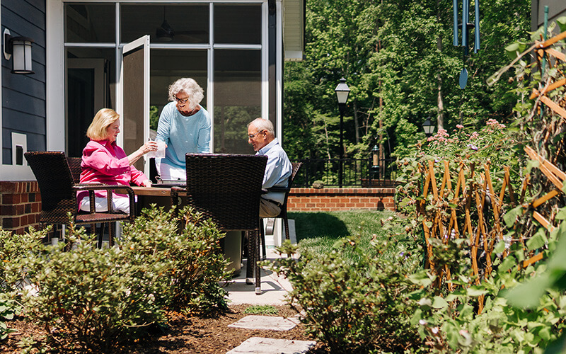 Residents Relaxing on a Sunny Patio Three seniors enjoying a sunny day on a patio with greenery at a community.