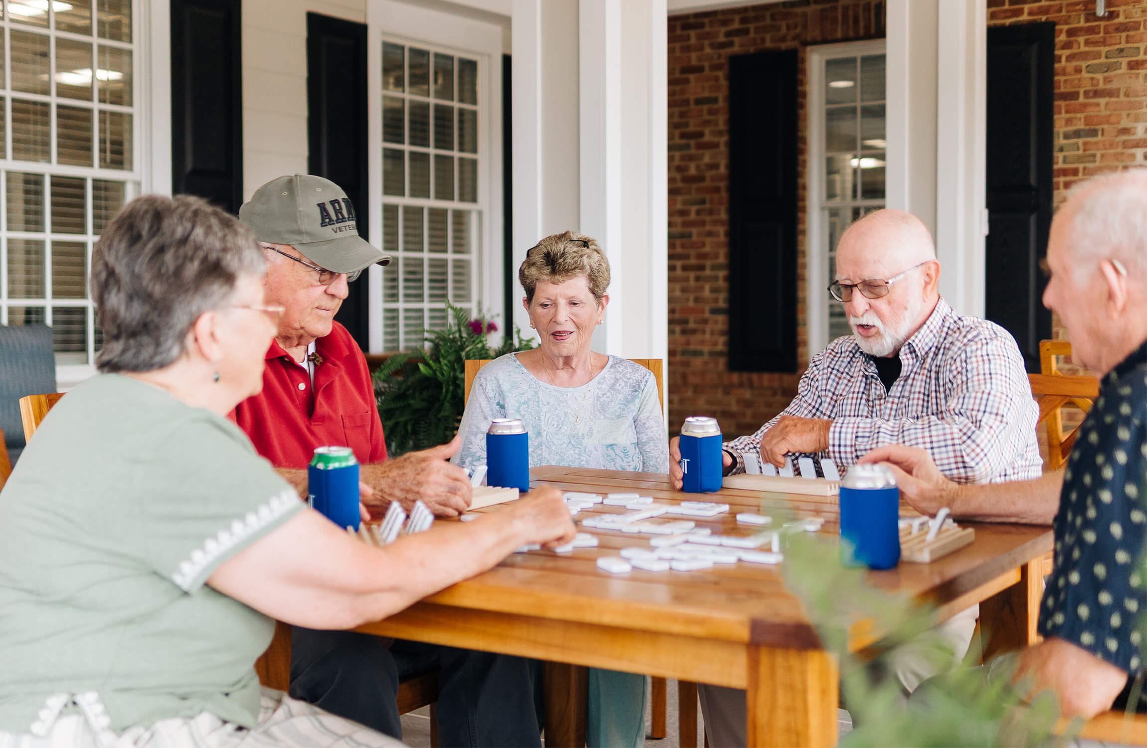 Group of seniors at a wooden table playing dominoes with drinks in hand.