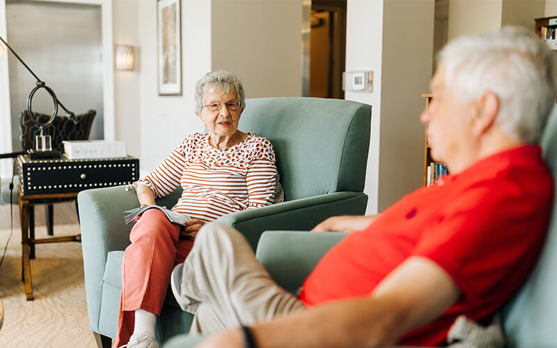 Two seniors relaxing in armchairs in a common area of their living community.