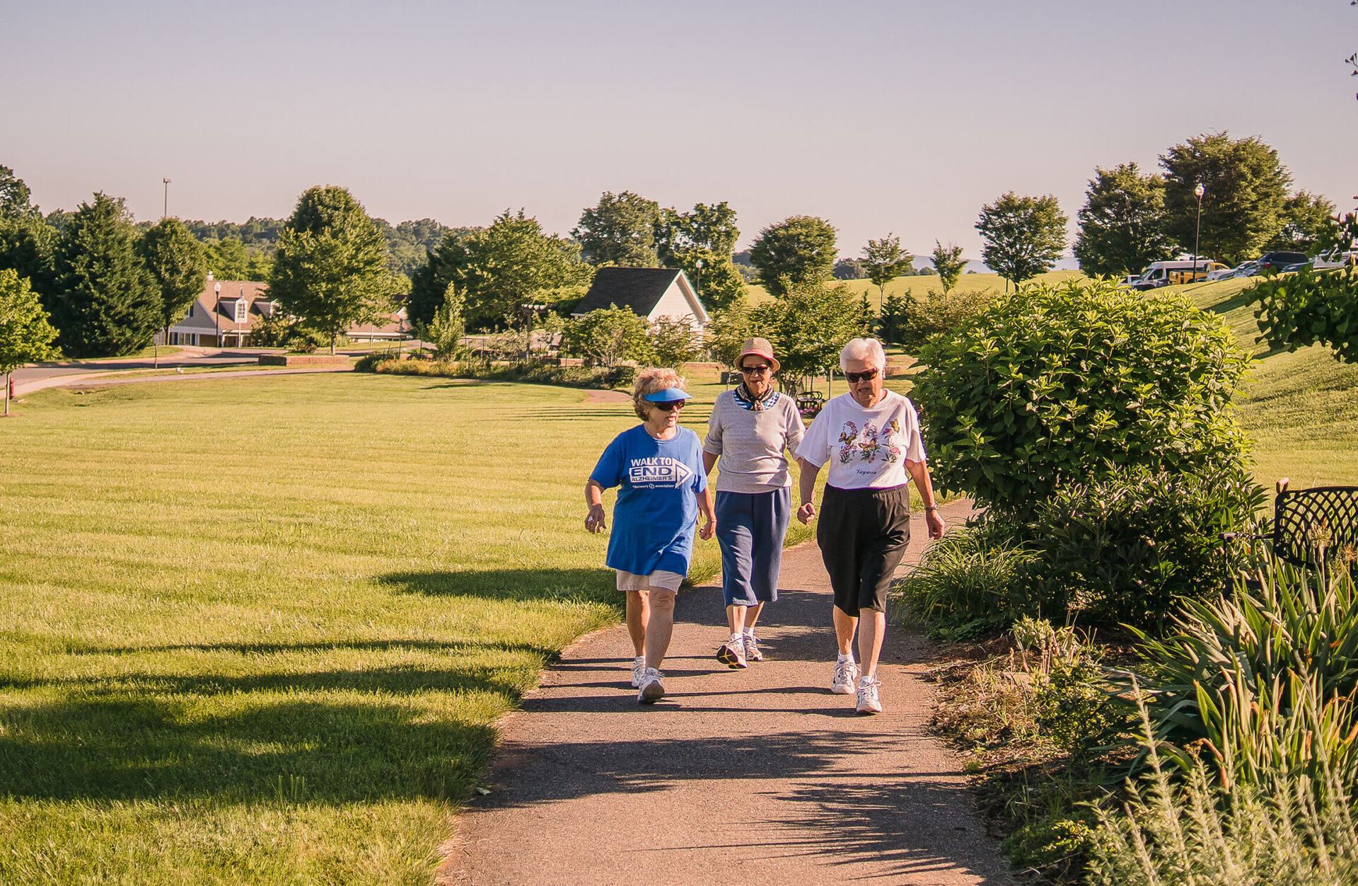 Three seniors enjoy a sunny walk on a garden pathway with units seen in the background.